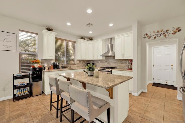 kitchen with stainless steel gas stove, a center island, white cabinets, and wall chimney range hood