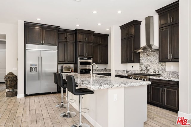 kitchen with sink, light hardwood / wood-style flooring, wall chimney exhaust hood, an island with sink, and stainless steel appliances