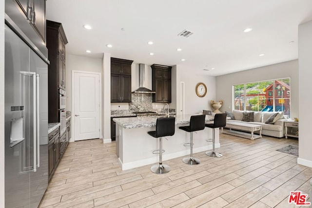 kitchen with a breakfast bar, a kitchen island with sink, wall chimney range hood, light hardwood / wood-style flooring, and light stone countertops