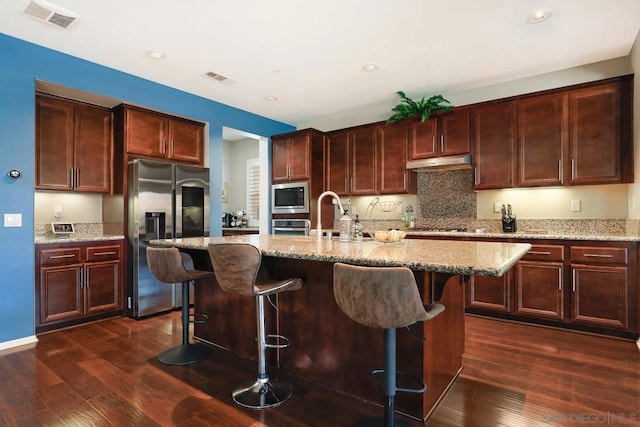 kitchen with a center island with sink, light stone counters, dark wood-type flooring, and appliances with stainless steel finishes