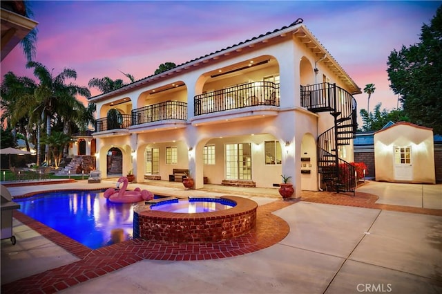 back house at dusk featuring a pool with hot tub, a patio area, and a balcony