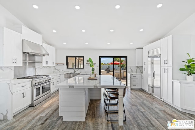 kitchen featuring light stone countertops, wall chimney exhaust hood, premium appliances, white cabinets, and hardwood / wood-style flooring