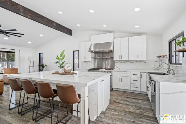 kitchen with a center island, wall chimney range hood, lofted ceiling with beams, a breakfast bar, and white cabinets