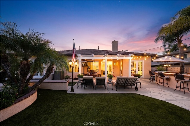 back house at dusk featuring a lawn, a bar, a patio, and an outdoor hangout area