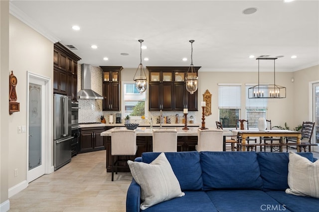 kitchen featuring decorative light fixtures, stainless steel appliances, a kitchen island with sink, and wall chimney range hood