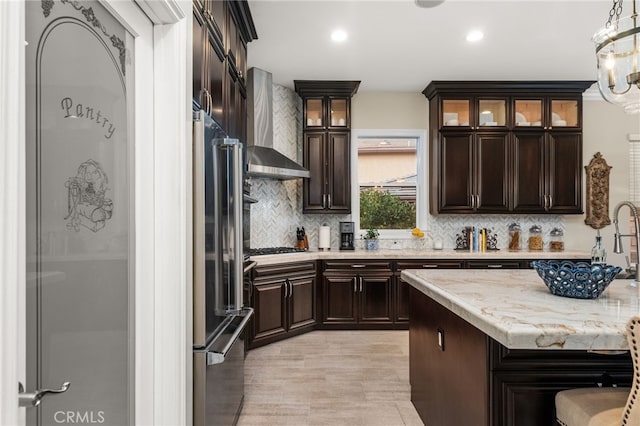 kitchen with hanging light fixtures, stainless steel appliances, wall chimney range hood, tasteful backsplash, and light hardwood / wood-style floors