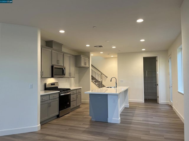 kitchen with sink, an island with sink, gray cabinets, appliances with stainless steel finishes, and hardwood / wood-style flooring