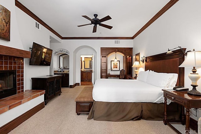 carpeted bedroom featuring a tiled fireplace, ornamental molding, ceiling fan, and ensuite bath