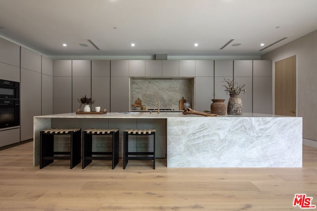 kitchen featuring a large island with sink, black double oven, light hardwood / wood-style floors, gray cabinets, and a breakfast bar