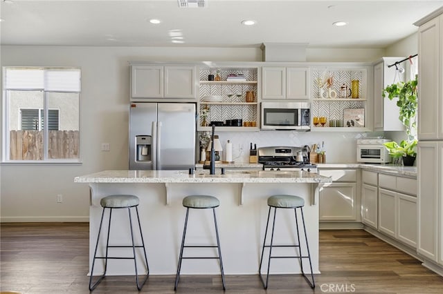 kitchen featuring stainless steel appliances, light stone counters, dark wood-type flooring, and an island with sink