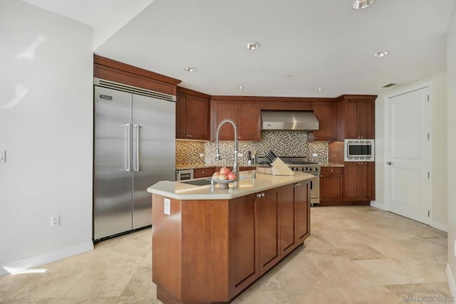 kitchen with sink, backsplash, built in appliances, an island with sink, and wall chimney exhaust hood