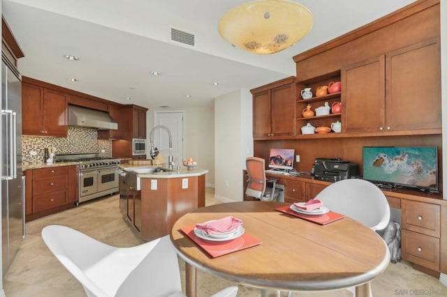 kitchen with wall chimney exhaust hood, sink, high end appliances, built in desk, and a kitchen island with sink