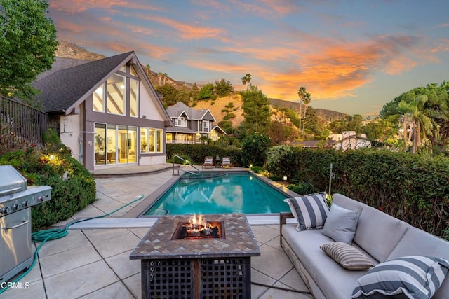 pool at dusk featuring a mountain view, a patio, area for grilling, and an outdoor living space with a fire pit