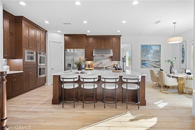 kitchen with decorative light fixtures, light wood-type flooring, an island with sink, and appliances with stainless steel finishes