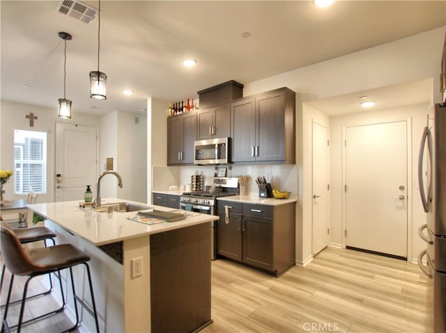 kitchen featuring stainless steel appliances, an island with sink, light hardwood / wood-style floors, decorative light fixtures, and dark brown cabinets