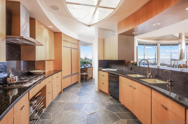 kitchen featuring dishwasher, stainless steel gas cooktop, light brown cabinetry, dark stone counters, and wall chimney exhaust hood