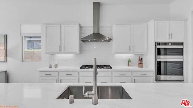 kitchen featuring wall chimney range hood, white cabinetry, stainless steel appliances, tasteful backsplash, and light stone countertops