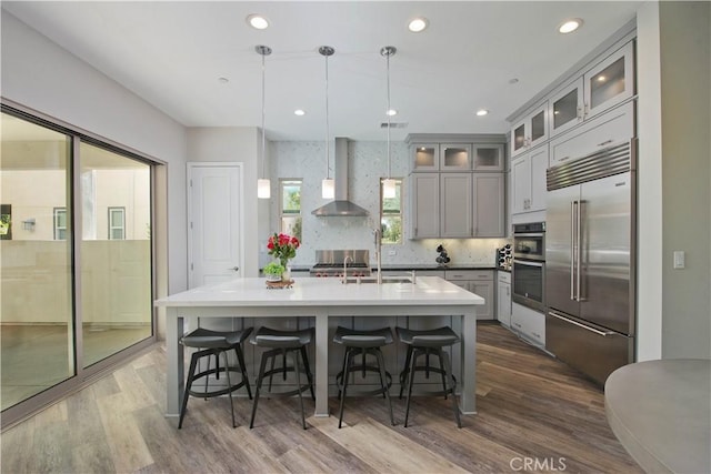 kitchen featuring wall chimney exhaust hood, stainless steel appliances, a kitchen island with sink, decorative light fixtures, and hardwood / wood-style floors