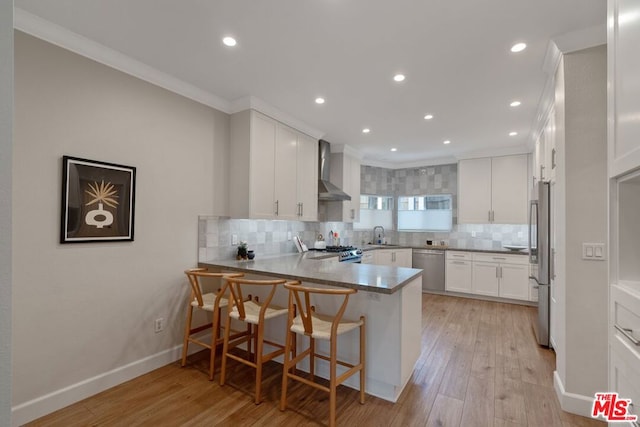 kitchen featuring white cabinetry, wall chimney exhaust hood, crown molding, light wood-type flooring, and appliances with stainless steel finishes