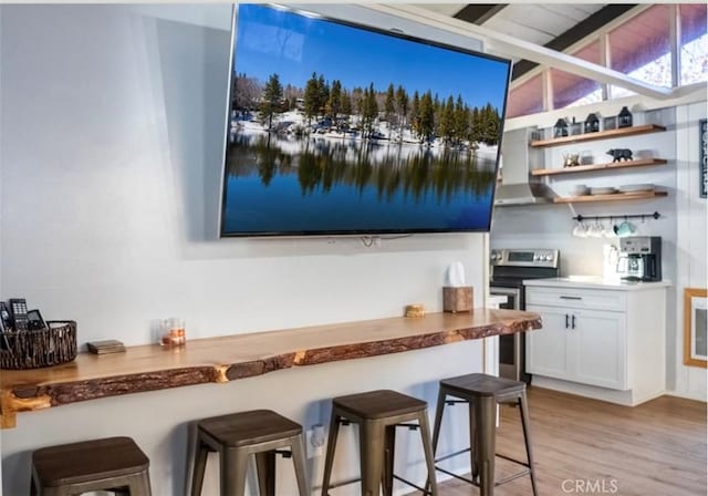 bar with wall chimney range hood, beamed ceiling, white cabinets, light hardwood / wood-style floors, and stainless steel electric range