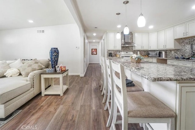 kitchen featuring a kitchen bar, light stone countertops, pendant lighting, white cabinets, and wall chimney range hood