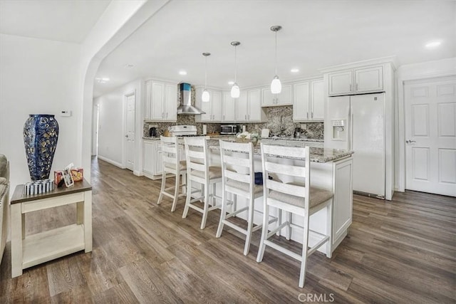 kitchen with decorative light fixtures, white cabinetry, light stone countertops, white refrigerator with ice dispenser, and wall chimney exhaust hood