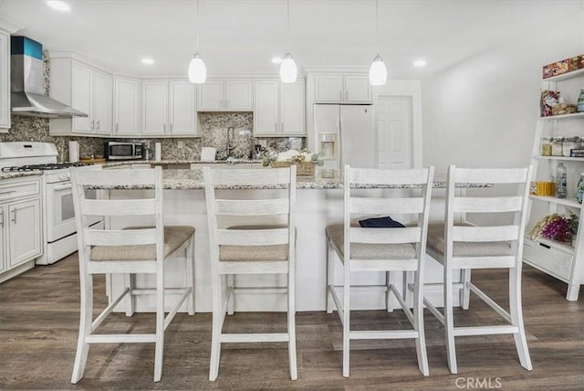 kitchen featuring pendant lighting, white cabinets, white appliances, wall chimney range hood, and a breakfast bar area