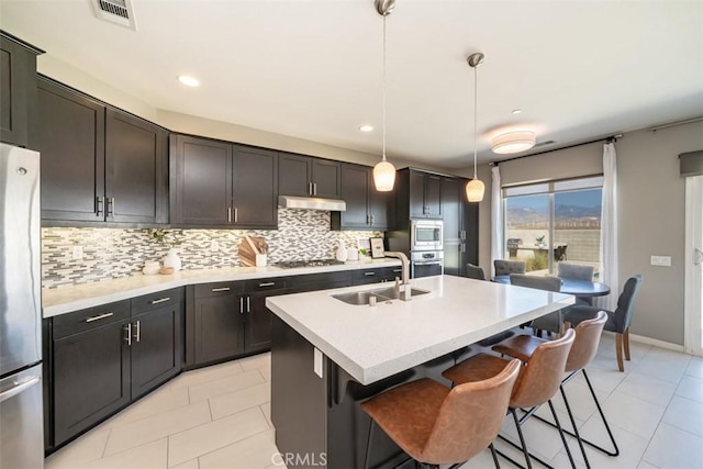 kitchen with sink, stainless steel appliances, hanging light fixtures, backsplash, and a kitchen island with sink