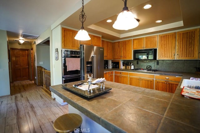 kitchen with a raised ceiling, tasteful backsplash, pendant lighting, light hardwood / wood-style floors, and black appliances