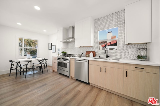 kitchen with light brown cabinetry, wall chimney exhaust hood, stainless steel appliances, sink, and light hardwood / wood-style flooring