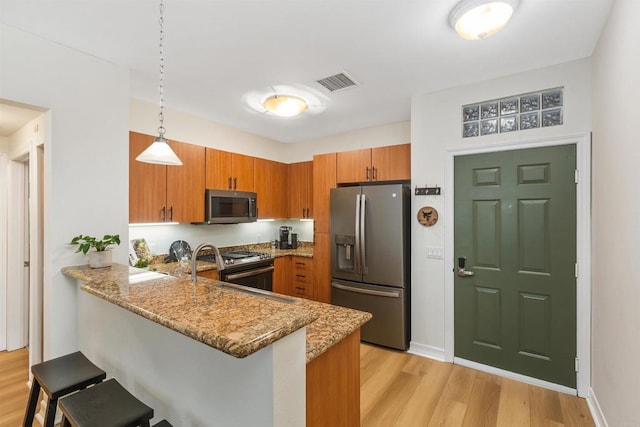 kitchen with brown cabinets, stainless steel appliances, light wood-style flooring, light stone countertops, and a peninsula