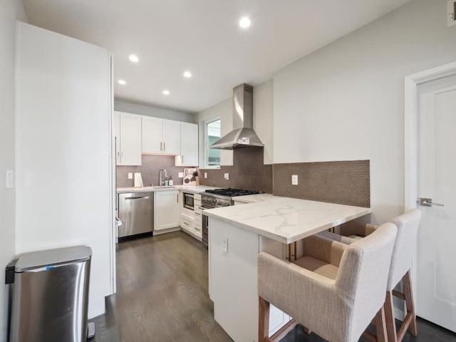 kitchen featuring a breakfast bar, white cabinets, wall chimney exhaust hood, kitchen peninsula, and stainless steel appliances