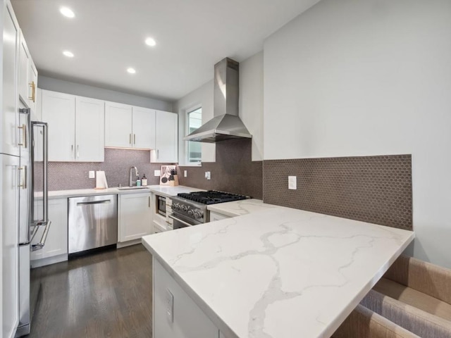 kitchen featuring light stone countertops, appliances with stainless steel finishes, kitchen peninsula, wall chimney exhaust hood, and white cabinetry