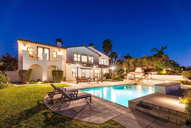 back house at night featuring a yard, an outdoor fireplace, a balcony, and a patio