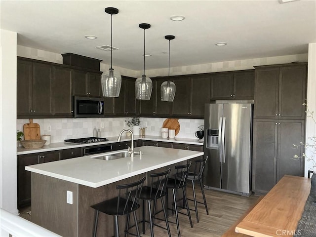 kitchen with sink, hanging light fixtures, light hardwood / wood-style floors, a kitchen island with sink, and appliances with stainless steel finishes