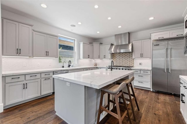 kitchen featuring wall chimney exhaust hood, light stone counters, appliances with stainless steel finishes, dark hardwood / wood-style floors, and an island with sink