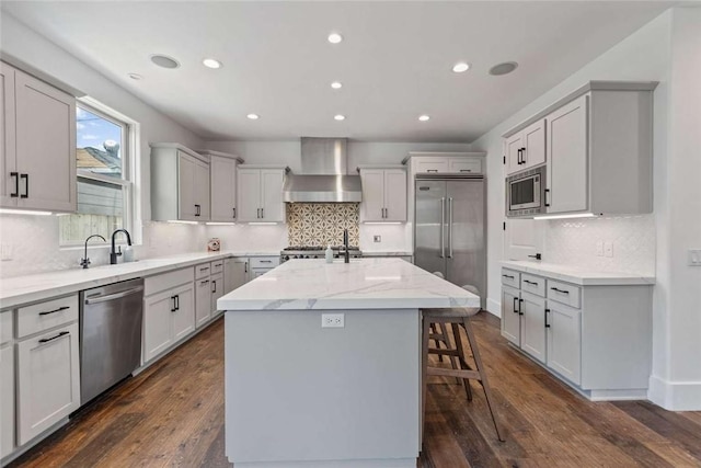 kitchen with a kitchen island with sink, built in appliances, light stone counters, dark wood-type flooring, and wall chimney range hood