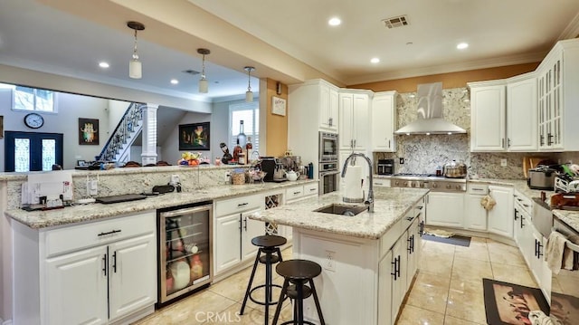 kitchen with a wealth of natural light, a kitchen island with sink, wall chimney exhaust hood, and beverage cooler