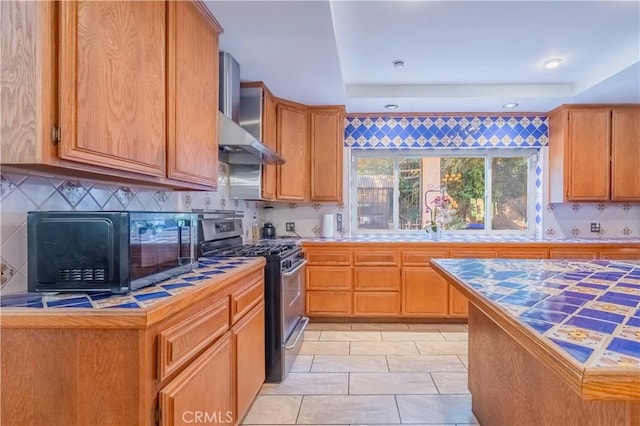 kitchen featuring backsplash, gas range, a tray ceiling, wall chimney range hood, and tile countertops