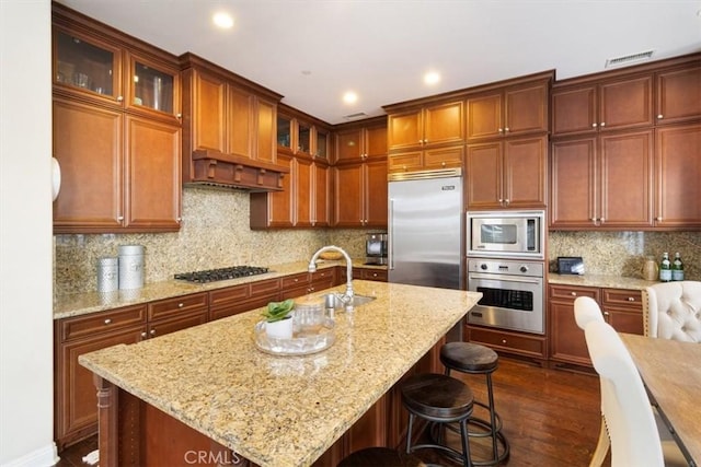 kitchen with built in appliances, dark hardwood / wood-style floors, backsplash, and an island with sink