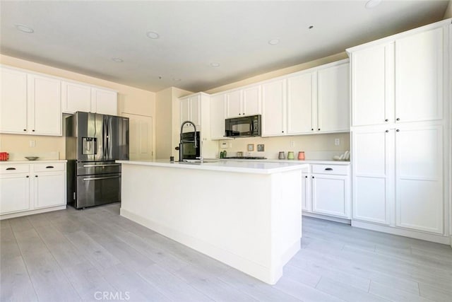 kitchen with light countertops, a kitchen island with sink, white cabinetry, black microwave, and stainless steel fridge