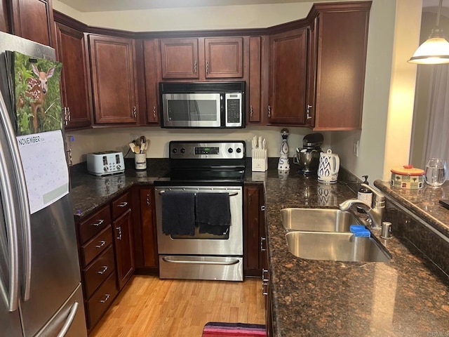 kitchen featuring appliances with stainless steel finishes, dark stone counters, light hardwood / wood-style floors, sink, and hanging light fixtures