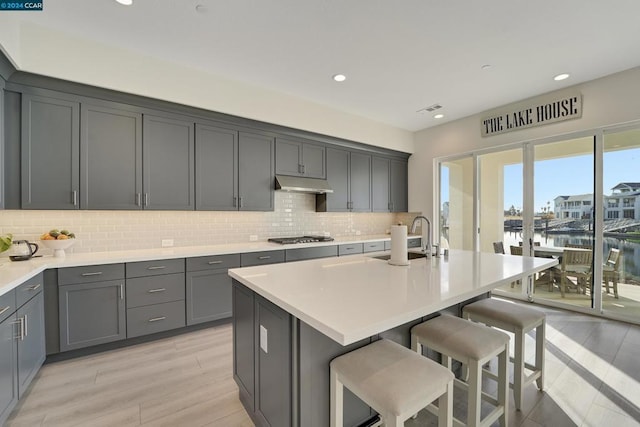 kitchen featuring light wood-type flooring, a kitchen island with sink, stainless steel gas cooktop, a water view, and gray cabinets