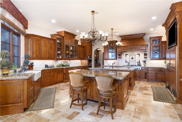 kitchen featuring stone tile flooring, a sink, custom exhaust hood, brown cabinetry, and an island with sink