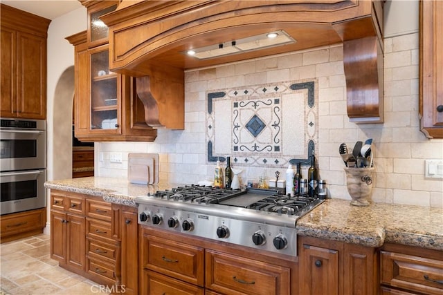 kitchen with glass insert cabinets, light stone countertops, decorative backsplash, brown cabinetry, and appliances with stainless steel finishes