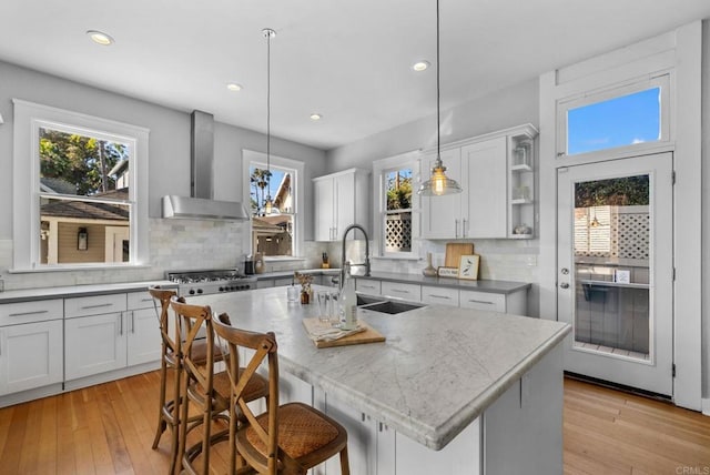 kitchen with wall chimney range hood, a kitchen breakfast bar, light hardwood / wood-style flooring, pendant lighting, and a kitchen island with sink