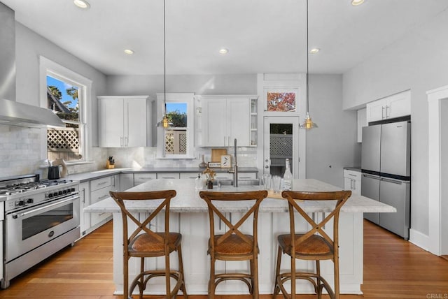 kitchen featuring wall chimney exhaust hood, an island with sink, pendant lighting, and appliances with stainless steel finishes