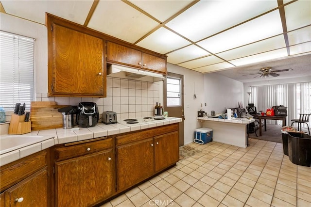 kitchen featuring light tile patterned floors, a ceiling fan, brown cabinetry, under cabinet range hood, and tasteful backsplash