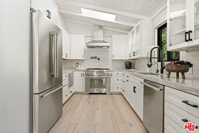 kitchen with high end appliances, white cabinets, sink, vaulted ceiling with skylight, and wall chimney exhaust hood