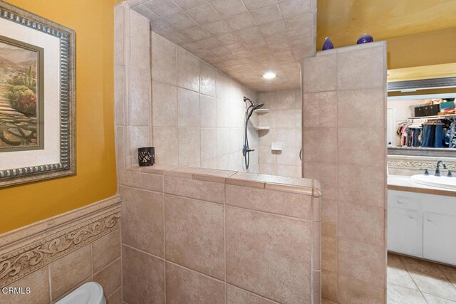 bathroom featuring a wainscoted wall, tile patterned flooring, a tile shower, vanity, and tile walls